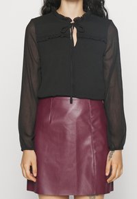 Svart blus med volangkrage och knytdetalj, matchad med en bordeauxfärgad läderkjol. Outfiten har långa genomskinliga ärmar och en figurnära silhuett.