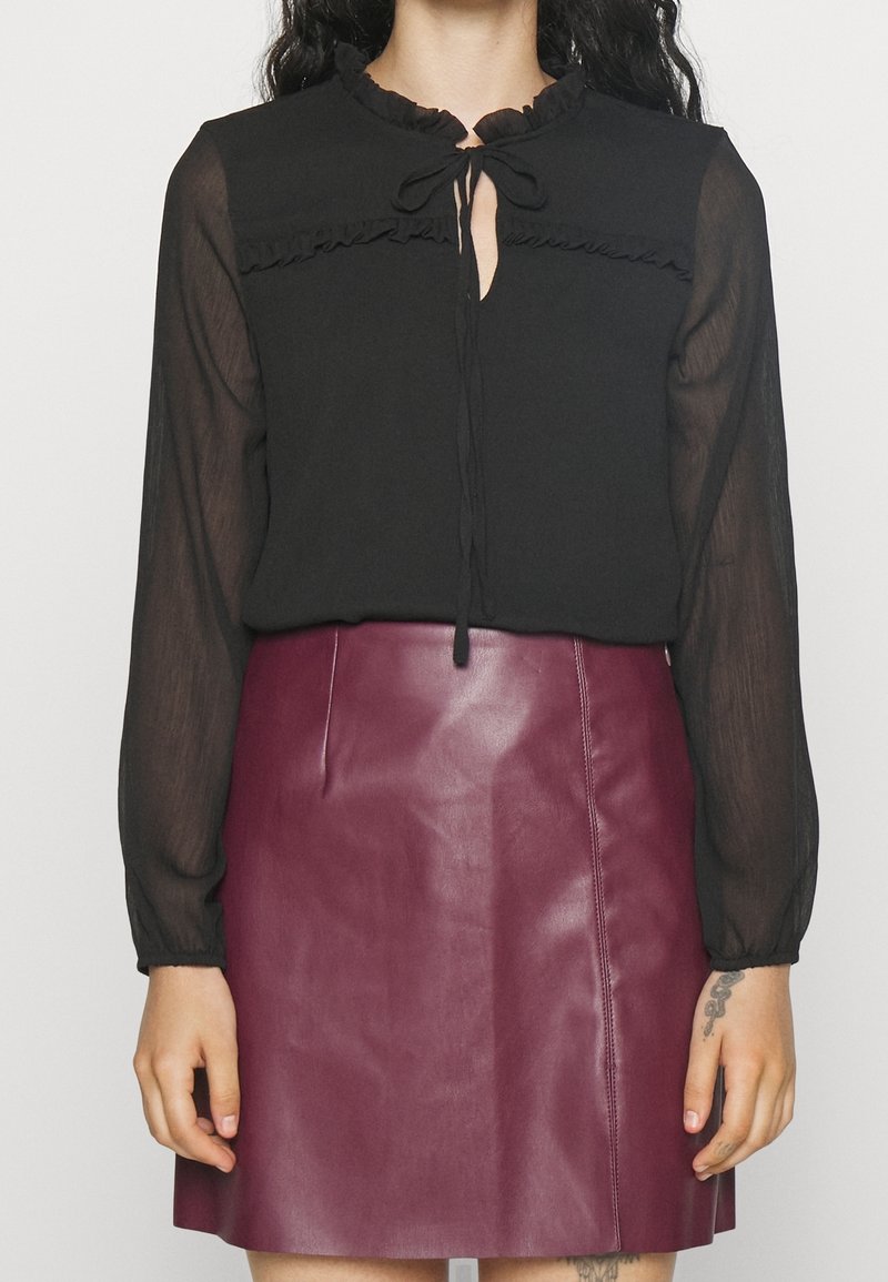 Svart blus med volangkrage och knytdetalj, matchad med en bordeauxfärgad läderkjol. Outfiten har långa genomskinliga ärmar och en figurnära silhuett.