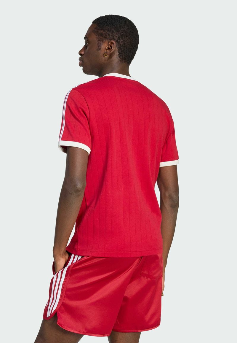 Homme de dos portant un t-shirt rouge côtelé à manches courtes avec un liseré blanc et un short rouge assorti avec des bandes blanches sur les côtés.