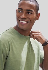 Green crew-neck t-shirt made of lightweight fabric, featuring a subtle texture. Model is adjusting the neckline, showing fitted sleeves.