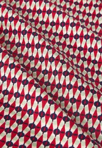 Zbliżenie złożonego materiału z powtarzającym się geometrycznym wzorem czerwonych diamentów, granatowych kół oraz jasnoszarych akcentów na białym tle.