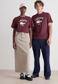 Vyno raudonos spalvos marškinėliai su "Lacoste" logotipu; béžiniai džinsiniai sijonai ir tamsiai m�ėlyni kelnės; juodi platforminiai batai; šviesiai rožinė kepurė; vienoda fono spalva.