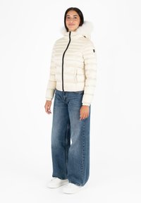 Giubbotto imbottito beige chiaro con collo alto e bordo in pelliccia sul cappuccio, chiusura a zip e polsini elastici. Abbinato a jeans blu a gamba larga e sneakers bianche.