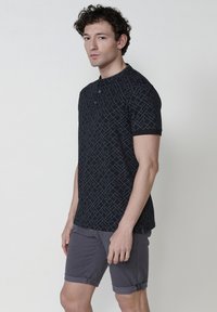 Crna polo majica s geometrijskim uzorkom u svijetloplavoj boji. Ima placket s tri gumba i kratke rukave. Uparen s sivim kratkim hlačama.