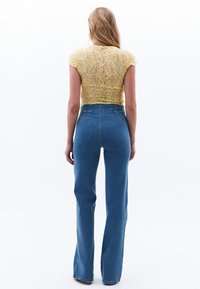 Blusa de encaje amarilla con mangas cortas combinada con jeans acampanados de tiro medio en denim azul. La vista trasera muestra la textura de la tela y el diseño de cintura alta.