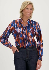 Multikleurige blouse met een abstract patroon in blauwe, oranje en roze tinten, met een striknek, lange mouwen en geplooide manchetten.