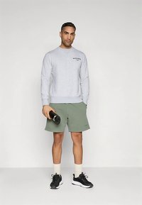 Sudadera gris claro con logo, combinada con pantalones cortos verde oliva. El modelo sostiene una botella de agua negra, lleva zapatillas negras y calcetines crema.