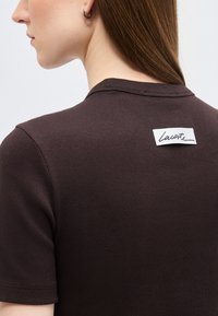 Femme portant un haut marron foncé côtelé à manches courtes avec une étiquette rectangulaire blanche portant l'inscription "Lacoste" dans le haut du dos.