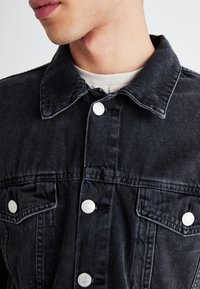 Donkerblauwe spijkerjasje met een klassieke kraag, witte knopen en twee borstzakken, gemaakt van een gestructureerde stof met subtiele stikdetails.