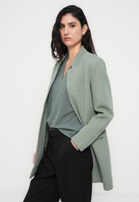 Πράσινο παλτό με ψαροκόκκαλο και μεσαία γιακά, μήκους πάνω από τους γοφούς, φορεμένο πάνω από ένα ταιριαστό τοπ. Μαύρο ψηλόμεσο παντελόνι ολοκληρώνει το σύνολο.