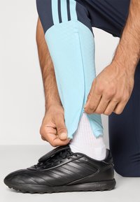 Χέρια προσαρμόζουν μια ανοιχτό μπλε προστατευτική κάλυψη μπάσου πάνω από λευκή κάλτσα και μαύρο παπούτσι στον λυγισμένο πόδι φορόντας ναυτικές παντελόνες με ανοιχτό μπλέ ρίγες.