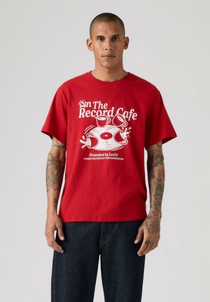 Hombre con tatuajes que lleva una camiseta roja con gráfico blanco y texto "On The Record Cafe", de pie frente a un fondo claro y liso.