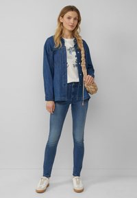 Veste en denim, t-shirt graphique blanc à fleurs, jeans skinny bleus, sac tressé beige et baskets crèmes avec semelles en gomme. Tenue décontractée.