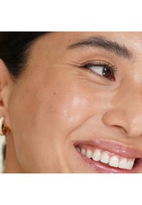 Primo piano del volto di una donna sorridente che mostra occhi marroni, lunghe ciglia, pelle liscia e labbra lucide, con un orecchino d'oro visibile.