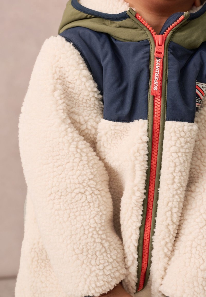 Enfant portant une veste sherpa polaire crème avec des panneaux d’épaule bleu marine et verts et une fermeture éclair rouge avec le logo Superdry.