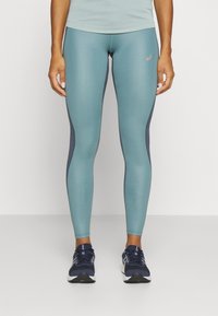 Lys blå leggings med mørkeblå sidelapper, tettsittende design, glatt tekstur og en liten logo på øvre venstre lår. Brukt med mørke sko.