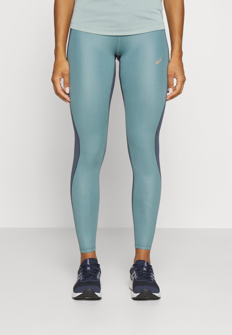 Lys blå leggings med mørkeblå sidelapper, tettsittende design, glatt tekstur og en liten logo på øvre venstre lår. Brukt med mørke sko.