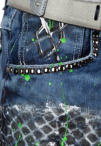 Jean en denim avec une base bleu foncé, des éclaboussures de peinture verte et des accents cloutés noirs le long de la poche, avec un design texturé et vieilli.