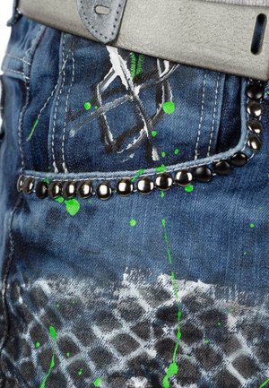 Jeans in denim con una base blu scuro, macchie di vernice verde e dettagli neri con borchie lungo la tasca, con un design testurizzato e consumato.