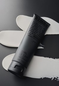 Tube noir de crème pour boucles avec texte en relief, posé sur des traces de crème blanche sur un fond sombre. Le capuchon du tube est gris et arrondi.