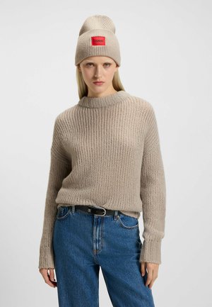 Beige gebreide trui met een ribbelstructuur, ronde hals en lange mouwen. Gepaarde met hoog-waisted blauwe jeans en een beige ribbel-beanie met een rode logopatch.