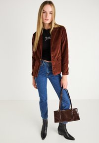 Brun fløjl blazer med to forlommer, sort grafisk cropped top, blå jeans og skinnende sorte ankelstøvler, holder en mørkebrun håndtaske.