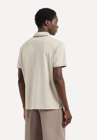 Camisa polo beige con collar acanalado y mangas cortas, que presenta un ribete negro a lo largo de los bordes, hecha de una tela suave y texturizada.