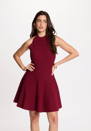 Frau mit langen dunklen Haaren, die ein ärmelloses, bordeauxfarbenes, geripptes Kleid mit tailliertem Schnitt trägt, steht mit den Händen in der Hüfte und schaut nach links vor weißem Hintergrund.