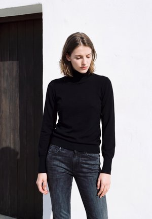 Une femme porte un pull noir à col roulé et un jean foncé, près d’une porte en bois sombre, devant un mur blanc.