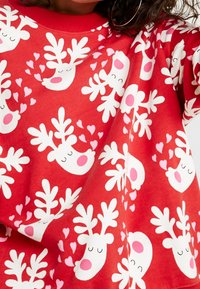 Sweat-shirt rouge avec un motif de renne blanc agrémenté d'accents roses, incluant des cœurs et des bois, en matériau doux et lisse.