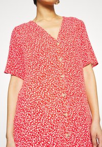 Femme portant une robe rouge à manches courtes avec imprimé floral blanc et boutons sur le devant, montrant le haut du corps et le cou.