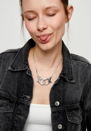 Mujer joven con chaqueta de mezclilla negra y camiseta blanca que lleva un collar grueso de cadena plateada, con los ojos cerrados y sacando la lengua de manera juguetona.