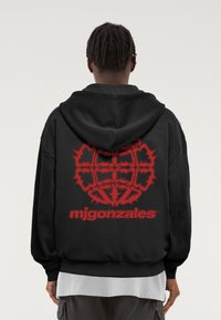 Fekete kapucnis pulóver, nagy piros grafikával a hátrészén, bonyolult mintákkal és az "mjgonzales" felirattal az alján.