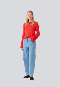 Red knit sweater with flared sleeves, paired with light blue high-waisted jeans. Model wears black slip-on shoes. Light gray background.
