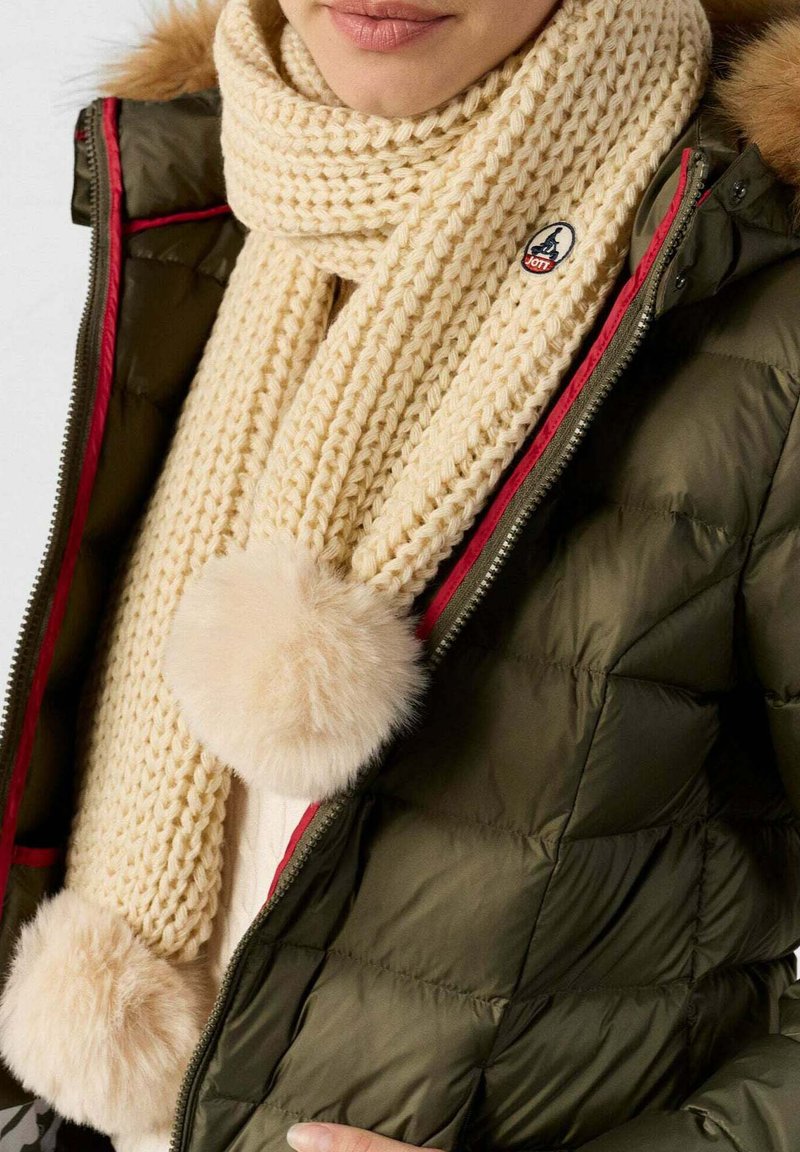 Beige gebreide sjaal met een gestructureerd patroon en grote bontbommels, gedragen over een donkergroene pufferjack met een rode rits.