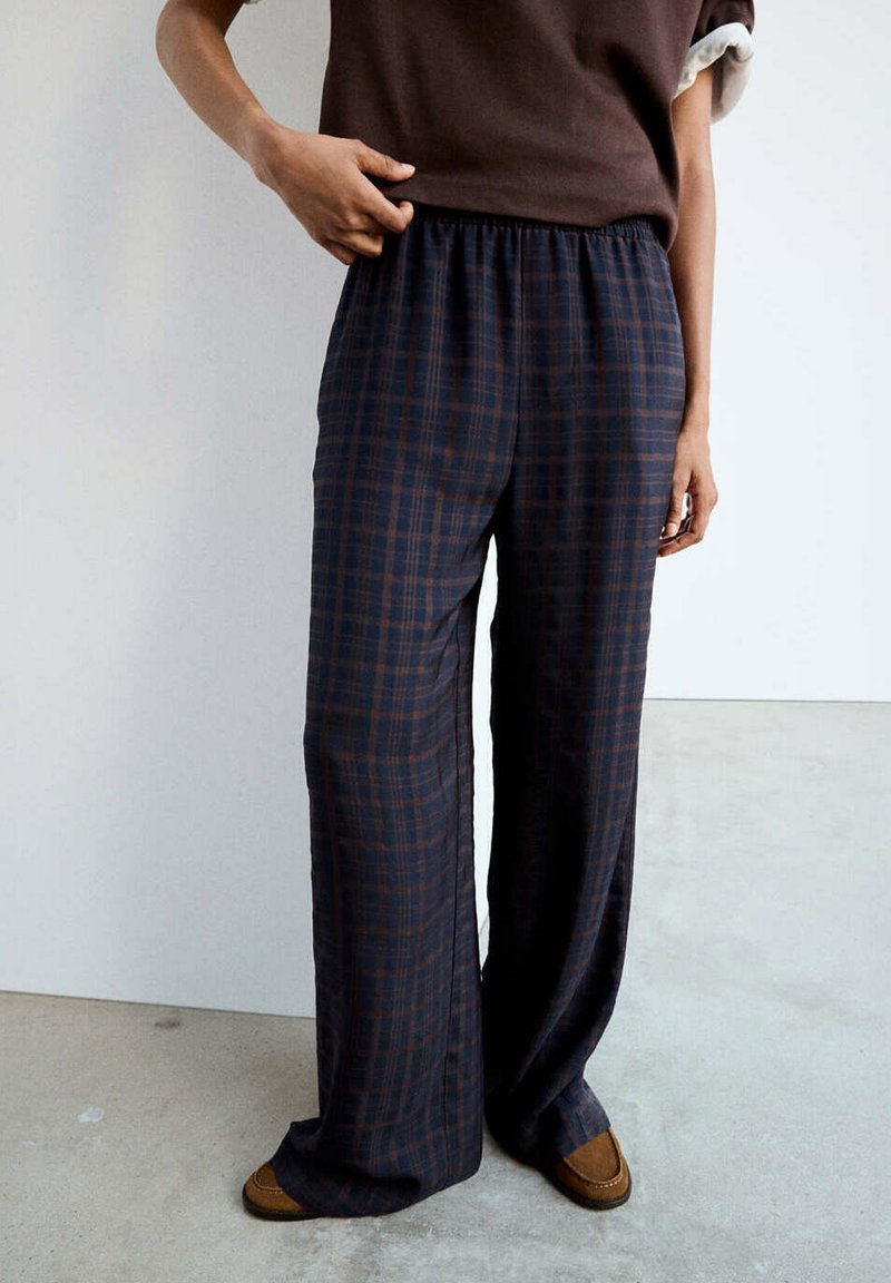 Personne portant un pantalon ample à carreaux foncés et des mocassins marron, ajustant la taille, debout sur un sol gris clair devant un mur blanc.