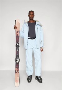 Άνδρας ντυμένος με ανοιχτό μπλε χιονοδρομική στολή και μαύρες χιονοδρομικές μπότες, κρατώντας ένα ζευγάρι ροζ και μαύρες σανίδες όρθιες.