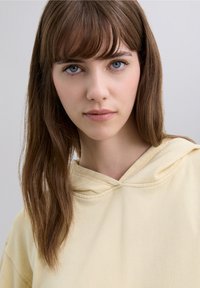 Hellgelber Kapuzenpullover mit lockerem Schnitt. Verfügt über eine gefütterte Kapuze und minimale Nähte, weiche Textur und gemütliches Aussehen.