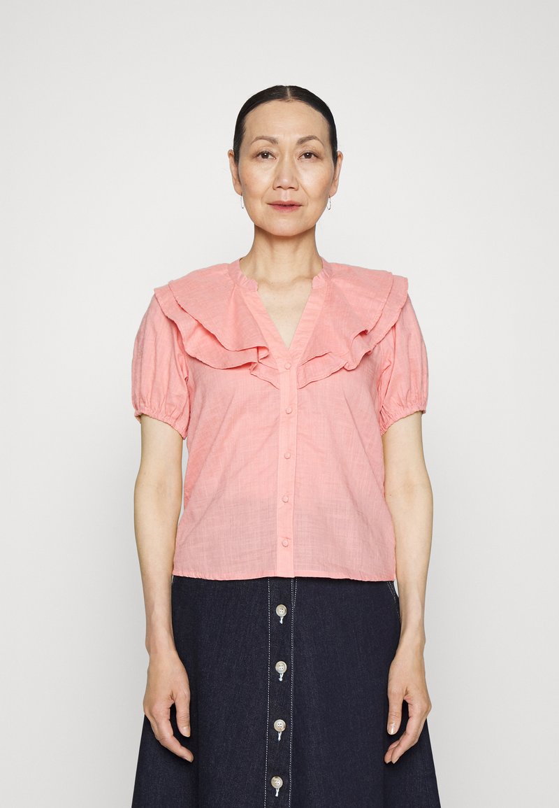 Blusa rosa com pregas, mangas curtas e bufantes, decote em V e frente com botões. Usada com uma saia de ganga escura com botões verticais.