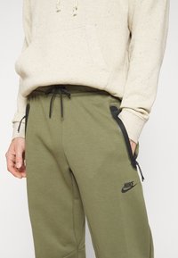 Oliwkowe dresowe spodnie z czarnymi akcentami i zamkami błyskawicznymi, z elastycznym paskiem w talii i sznurkiem do regulacji. Logo Nike nadrukowane z boku.