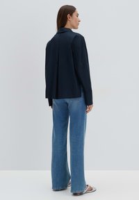 Camicia button-up blu navy con maniche lunghe, una piega sul retro e spalle strutturate, indossata sopra jeans larghi blu chiaro.