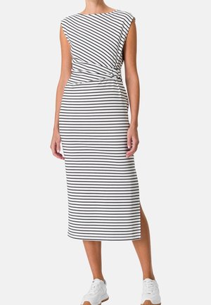 Vestido midi blanco sin mangas con rayas horizontales negras y abertura lateral, llevado con zapatillas deportivas blancas por una mujer de pie.