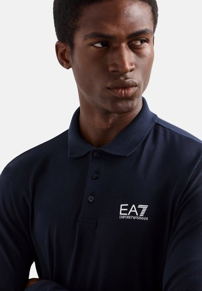 Jeune homme portant un polo bleu marine avec le logo EA7 Emporio Armani, regardant de côté sur un fond clair uni.
