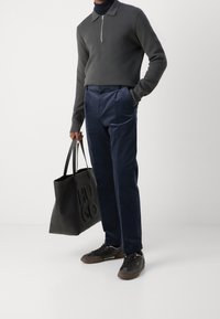 Grå långärmad zip-up tröja, marinblå kordbyxor, svart tote-väska med präglad logotyp och mörka sneakers med bruna accenter.