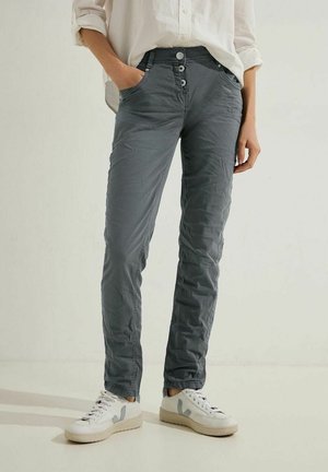 Frau trägt zerknitterte graue Hose, weiße Sneaker mit grauen "V"-Logos und ein weißes langärmliges Hemd mit hochgekrempelten Ärmeln, steht mit den Händen in den Taschen.