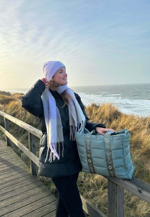 Polsternde türkisfarbene Tragetasche mit grauen Trägern, auf denen der Text "HEY MARLY" zu sehen ist; eine Frau in einem schwarzen Mantel, mit einem hellen Schal und einem lavendelfarbenen Hut steht auf einem Holzsteg.