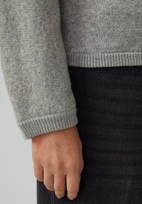 Grauer Strickpullover mit gerippten Bündchen, der eine weiche Textur zeigt. Hand vor einem schwarzen Stoffhintergrund platziert.