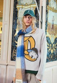 Pletena jopica v bež barvi z velikim grafičnim motivom Donalda Ducka, v kompletu z večbarvnim, prugastim volnenim šalom v modri in svetlo rjavi barvi.