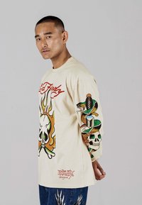 Beige Langarmhemd mit auffälligen Grafiken eines Schädels, Flammen und einer Schlange. Das Material wirkt weich; das Design ist lebhaft mit unterschiedlichen Farben.