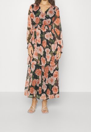 Kvinde iført en lang sort kjole med store pink og ferskenfarvede roser, gennemsigtige lange ærmer og beige høje hæle med remme.